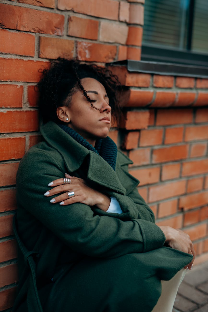 woman in green trench coat sitting and leaning against a concrete red brick wall