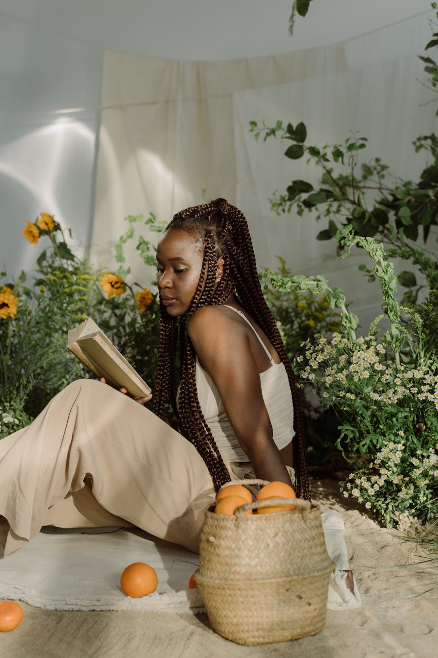 a woman with braided hair sitting on the picnic blanket reading a book